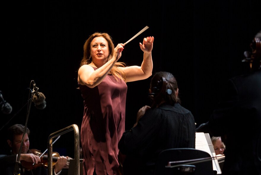 Concert conductor Simone Young AM, conducting on stage in a plum-coloured dress, musicians around her.