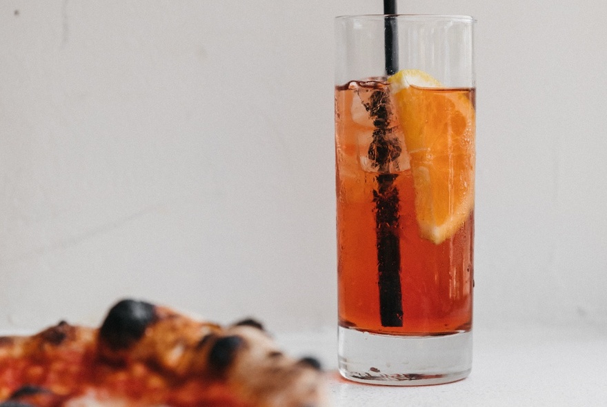 A high-ball glass of Aperol Spritz with a black straw and piece of orange as garnish, next to an out-of-focus pizza.