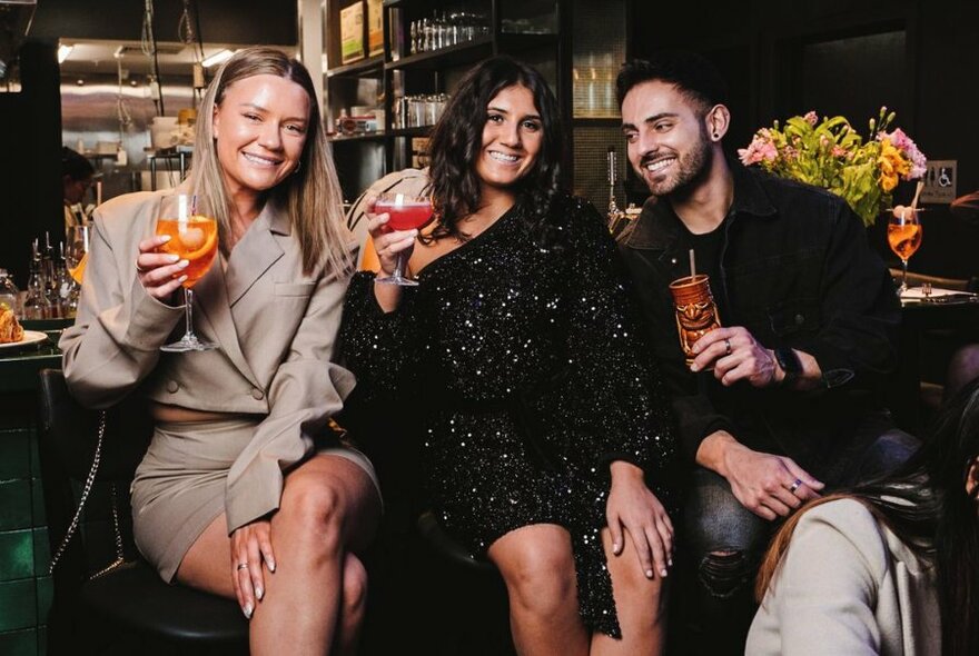 Three people sitting at a bar holding large cocktails.