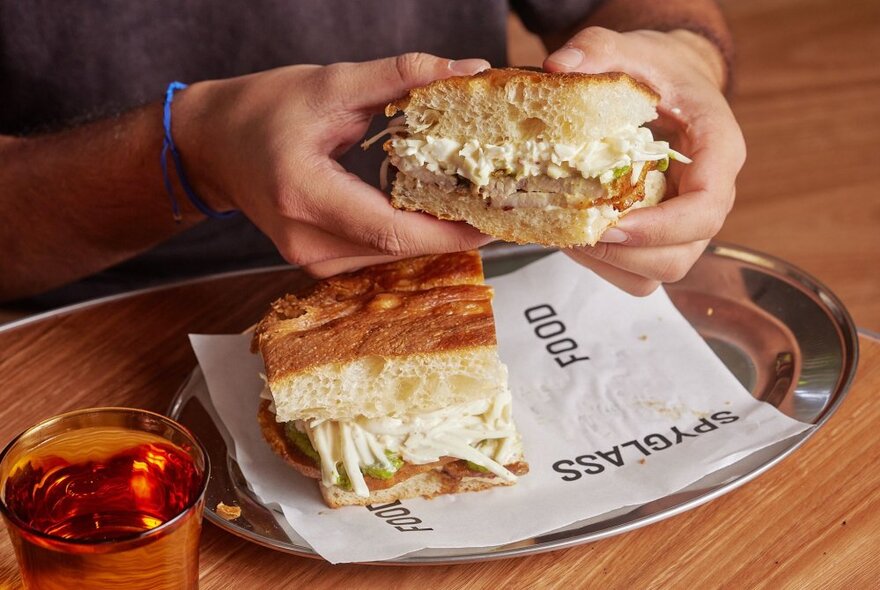 Hands holding large foccacia sandwich over a plate with napkin and drink.