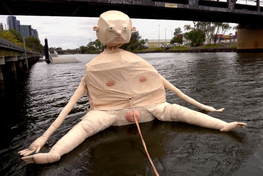 Naked baby sculpture floating on water near a bridge.