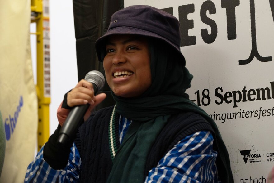 A person speaking into a microphone at an event in front of a partially obscured banner for the Emerging Writers' Festival.