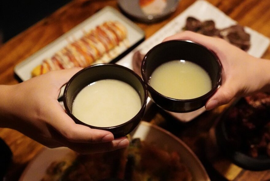 Two hands each holding a small bowl of a creamy Korean soup over a table laid with other Korean delicacies. 