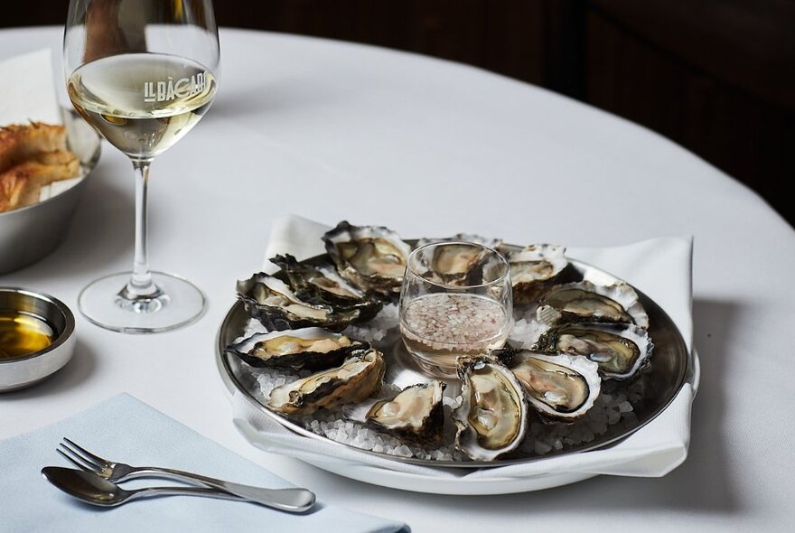 A platter of freshly shucked oysters on ice, on a white linen table top, with a glass of wine and cutlery on the table.