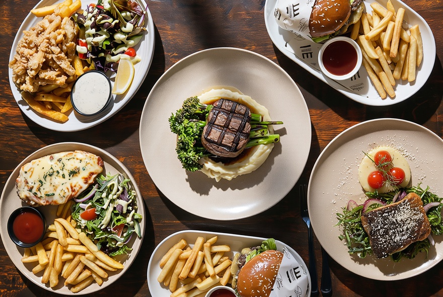 Looking down at plates of food that include steaks, burgers, salads and fries on a wooden restaurant dining table.