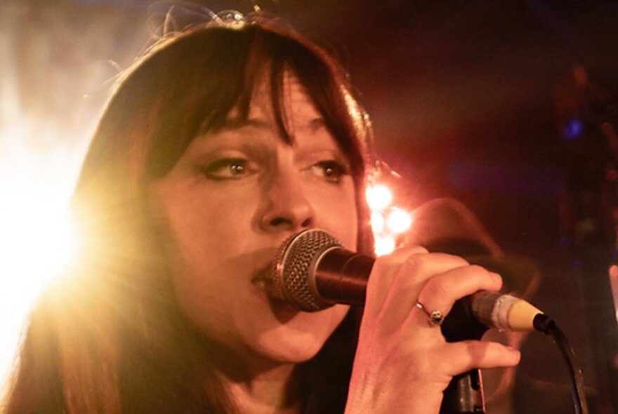 Singer Susy Hull singing into a microphone with a bright light behind her.