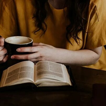 The body of a woman in a yellow t-shirt holding a mug reading an open book.