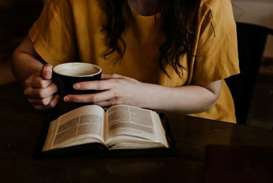 The body of a woman in a yellow t-shirt holding a mug reading an open book.
