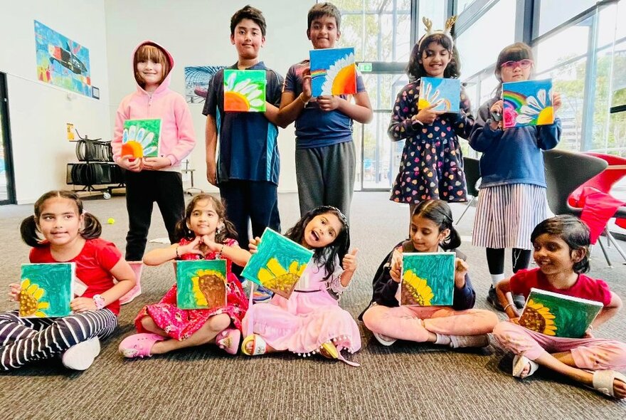 A standing and seated row of children in a large bright room, all holding up artworks they've created.