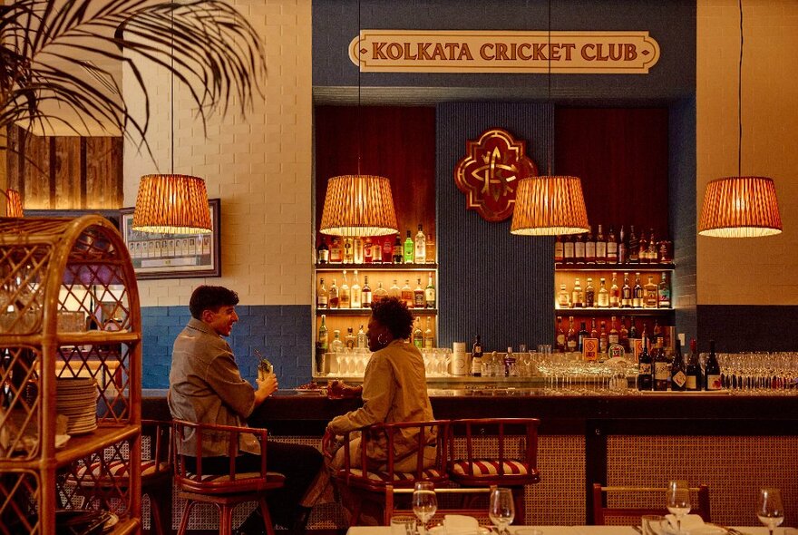 Two people facing each other seated at the bar of the Kolkata Cricket Club, pale orange light shades hanging from the ceiling, dining tables in the foreground.