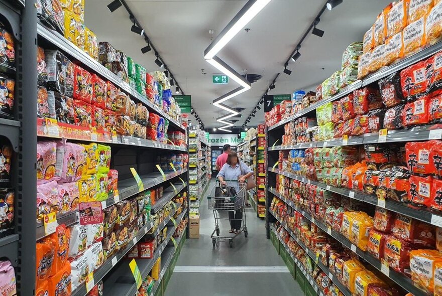 People pushing a trolley between aisles of shelves in a supermarket lit by modern jagged lighting, with rows of brightly coloured packaged food.