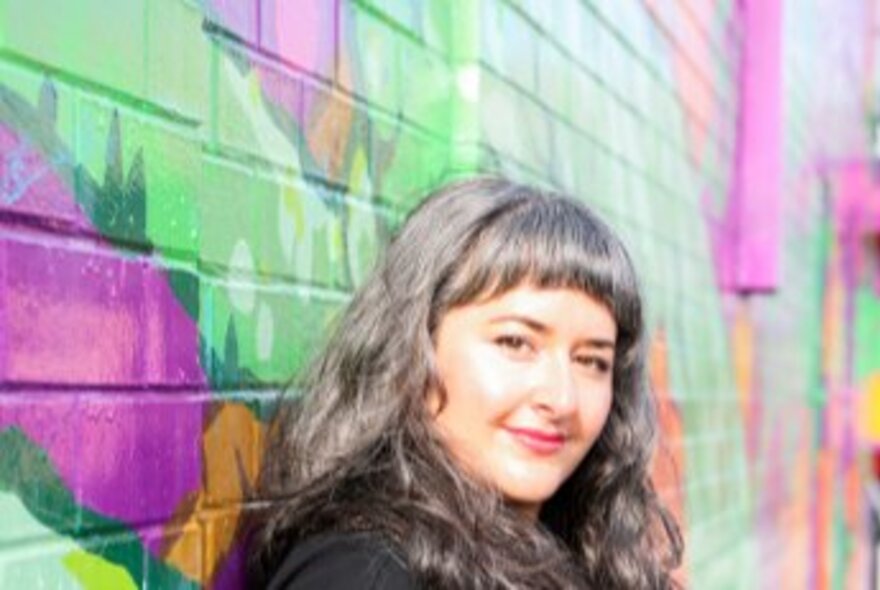 Author Alice Robinson, a person with long straight brown hair and a fringe, leaning against a brightly painted wall outdoors, sunlight on their face.