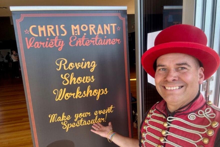 Christ Morant wearing a red ringmaster's coat and hat standing in front of a sign for his roving shows and workshops. 
