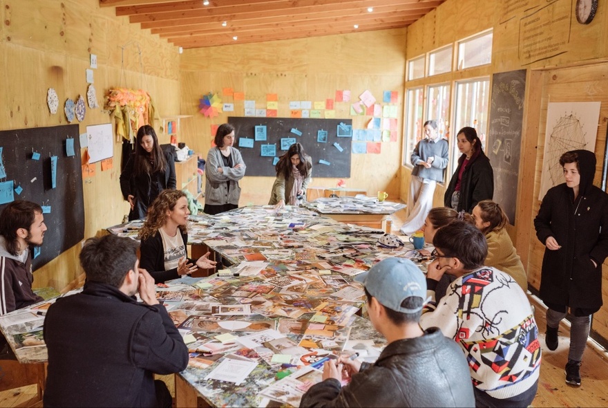 Participants in a creative workshop working at a table covered in artwork, in a room with walls covered in notes and artwork.