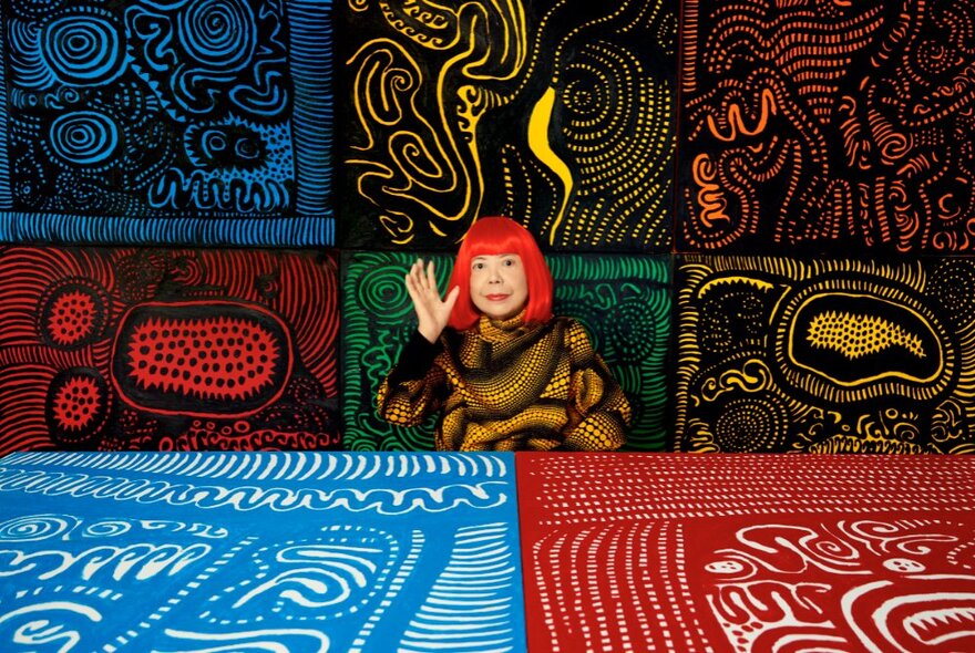 The artist Yayoi Kusama, with a bright red bob hairstyle, seated in front of a selection of her painted artworks displayed on a wall behind her and a table in front of her. 