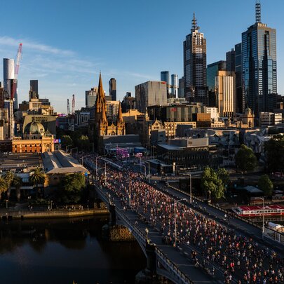 A large group of people taking part in the Nike Melbourne Marathon Festival, running across Princess Bridge over the Yarra River, with tall city buildings and trees in the background, set against a blue sky.
