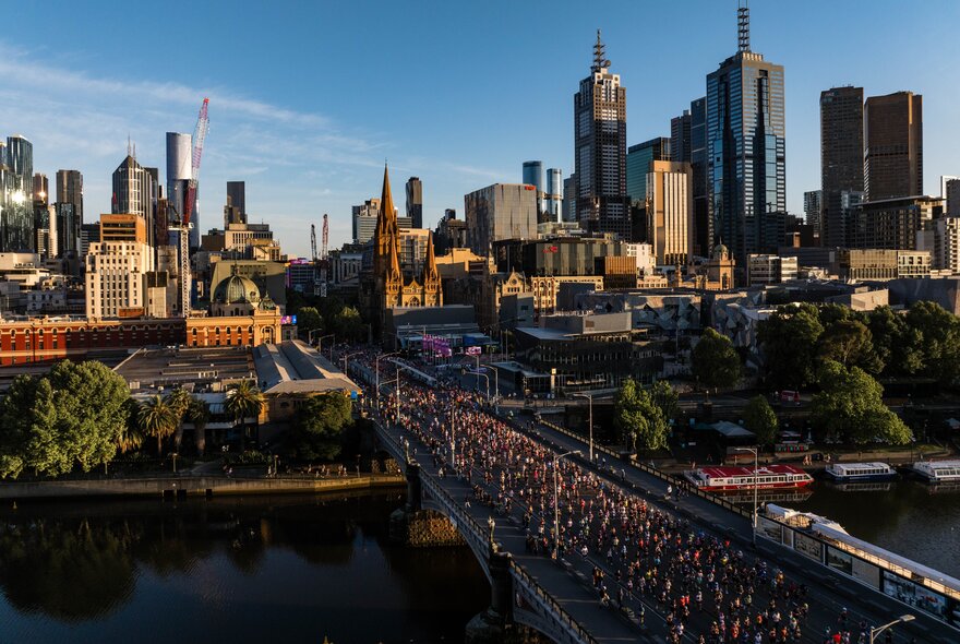 A large group of people taking part in the Nike Melbourne Marathon Festival, running across Princess Bridge over the Yarra River, with tall city buildings and trees in the background, set against a blue sky.