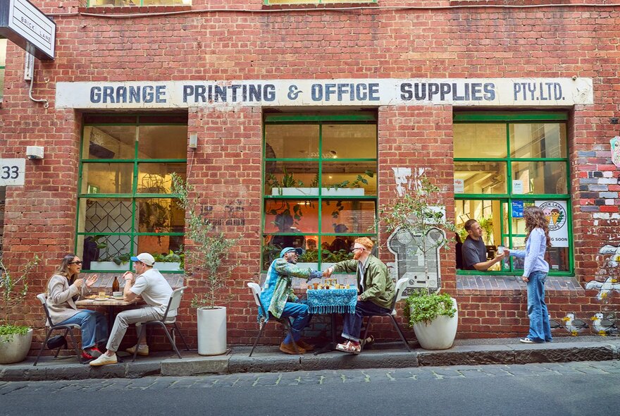 People dining outside a brick building in a laneway. A woman is ordering a coffee through a takeaway window.