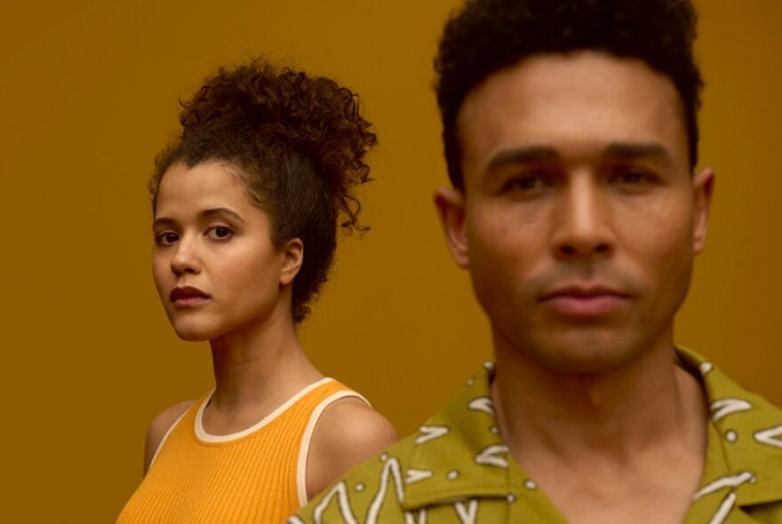 Two actors, male actor to the front with short dark hair and green shirt, female actor standing to the rear with a yellow tank top and curly hair piled on top of her head.