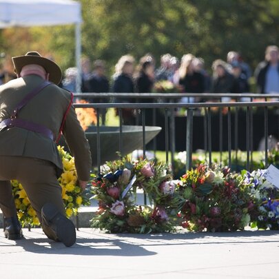 A person in army uniform kneeling and laying a wreath of flowers where other flowers have been placed. 