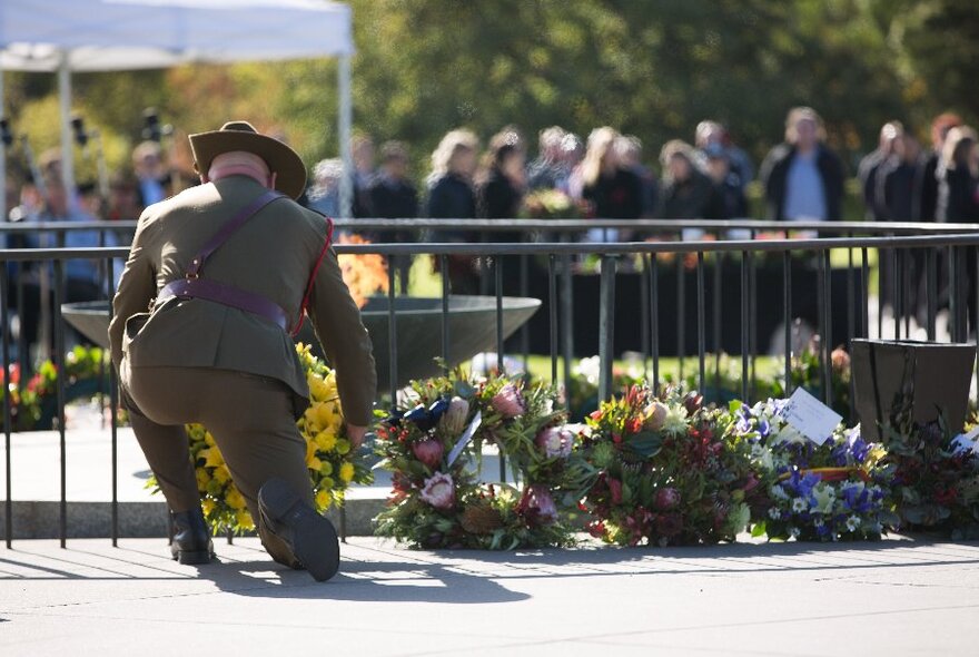 A person in army uniform kneeling and laying a wreath of flowers where other flowers have been placed. 