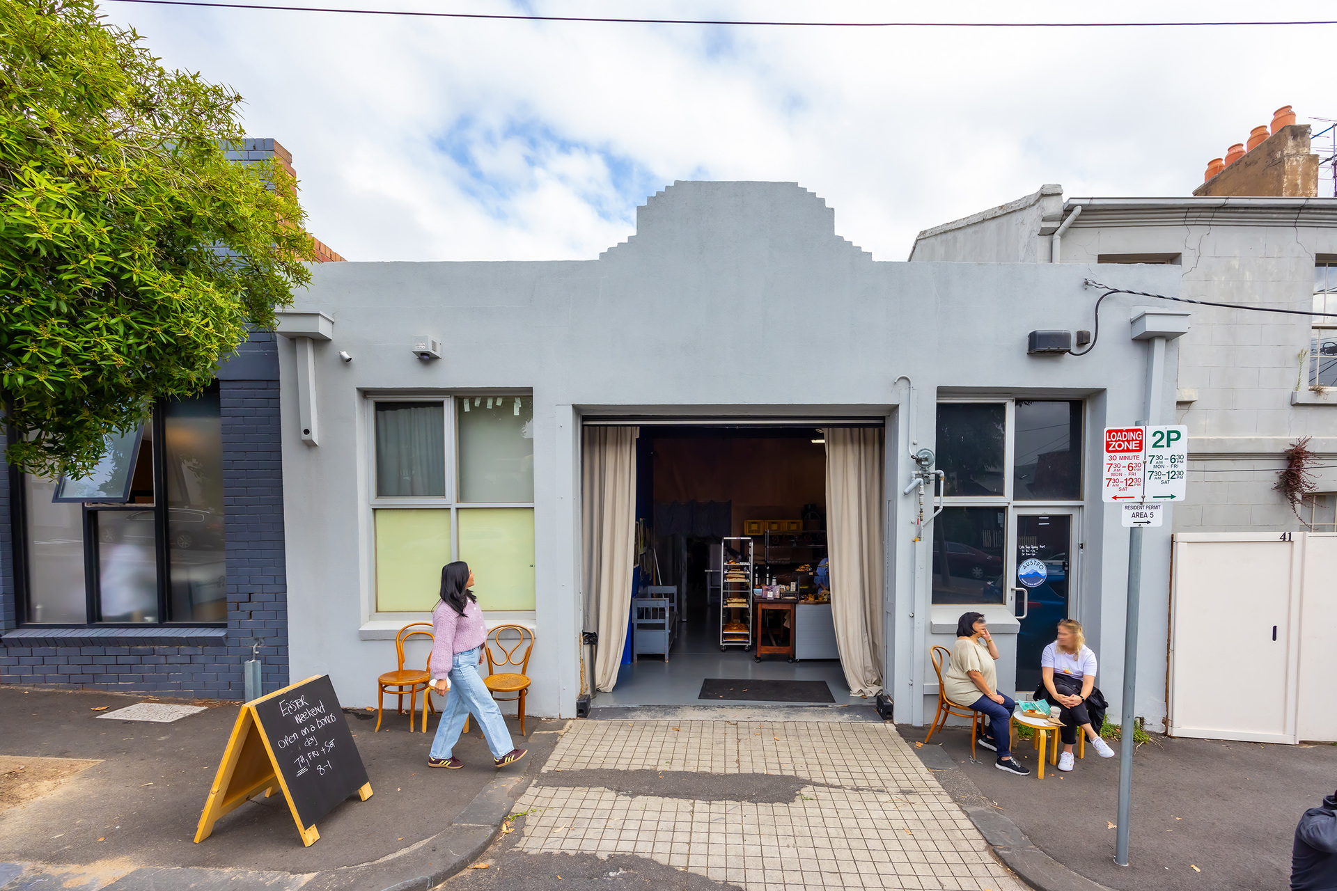 A person is walking past a bakery there are two people seated outside 