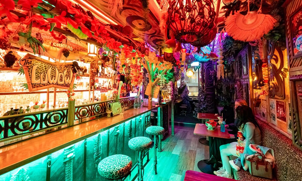 Two friends are drinking cocktails inside a kitschy tiki bar covered in tropical decorations.