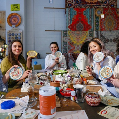 A group of people seated at a craft table covered in pots of paint and craft accessories, each holding their painted bowl.