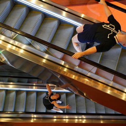 Two people bending backwards in a dance move on separate moving escalaters seen from above.
