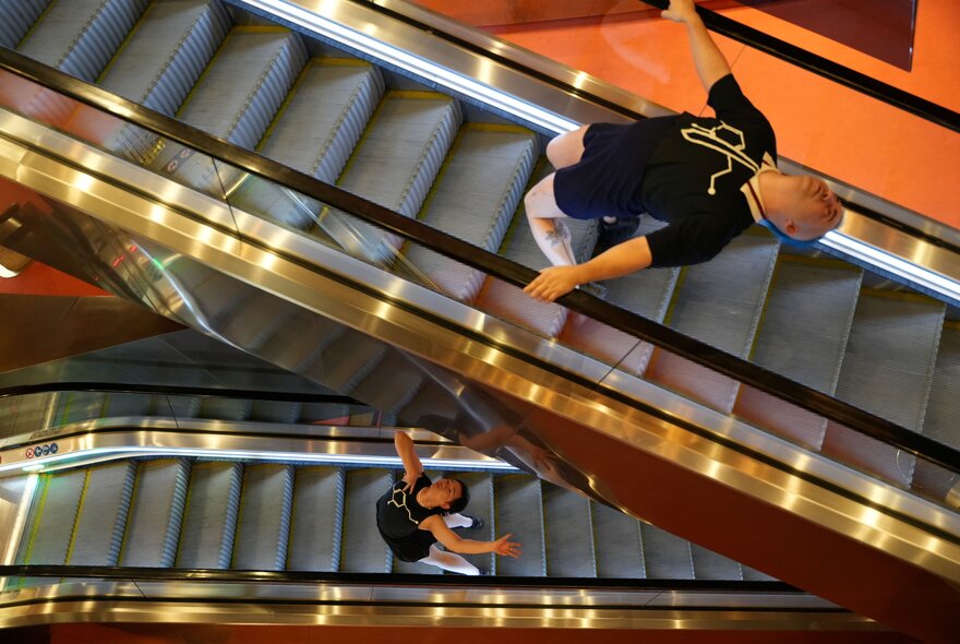 Two people bending backwards in a dance move on separate moving escalaters seen from above.