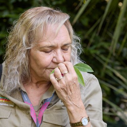 A woman in park ranger attire closes her eyes and sniffs a leave in a garden. 