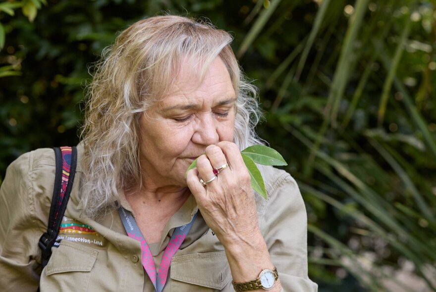 A woman in park ranger attire closes her eyes and sniffs a leave in a garden. 