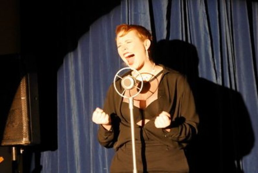 An impro performer on a stage singing expressively into a microphone, their fists clenched.