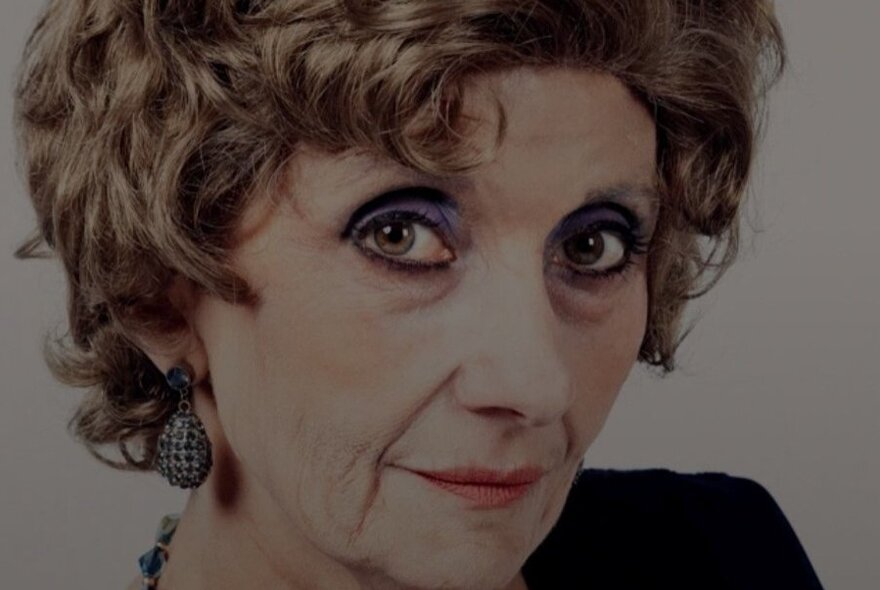 Close-up portrait of a woman with short, curly brown hair. She is wearing heavy, dark eye makeup.