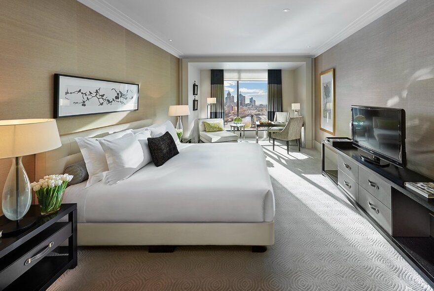 Hotel room with white king-sized bed facing a TV console with lamp and flowers, leading to seating area and floor to ceiling window with city views.