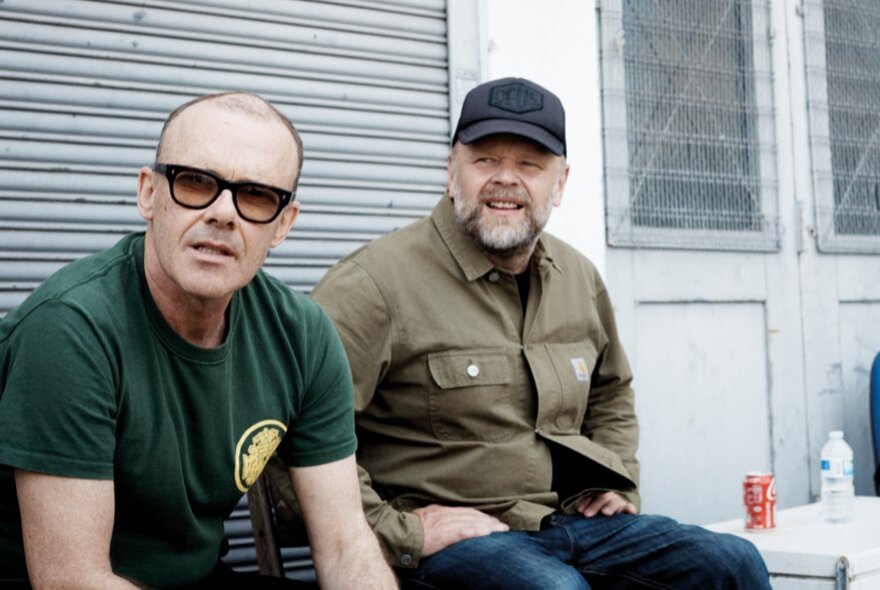 The two middle-aged men of Leftfield, seated in front of a garage roller door, one wearing a green t-shirt and sunglasses, the other wearing a khaki jacket and cap.
