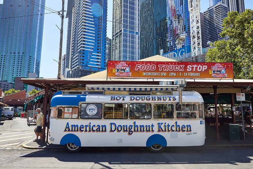 A white and blue van with hot doughnuts wording parked in front of a city market.