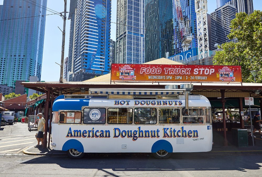 A white and blue van with hot doughnuts signage parked in front of a city market.