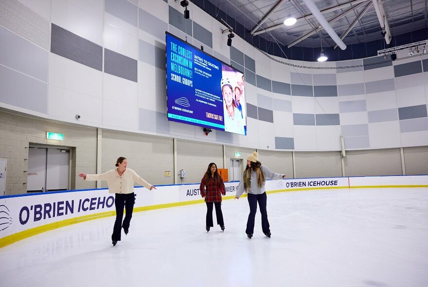 Three girls ice skating on an indoor ice rink.