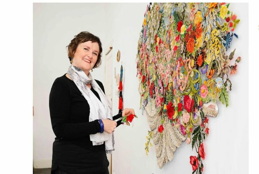 A woman standing next to a colourful, large wall hanging artwork made of wool and textiles.