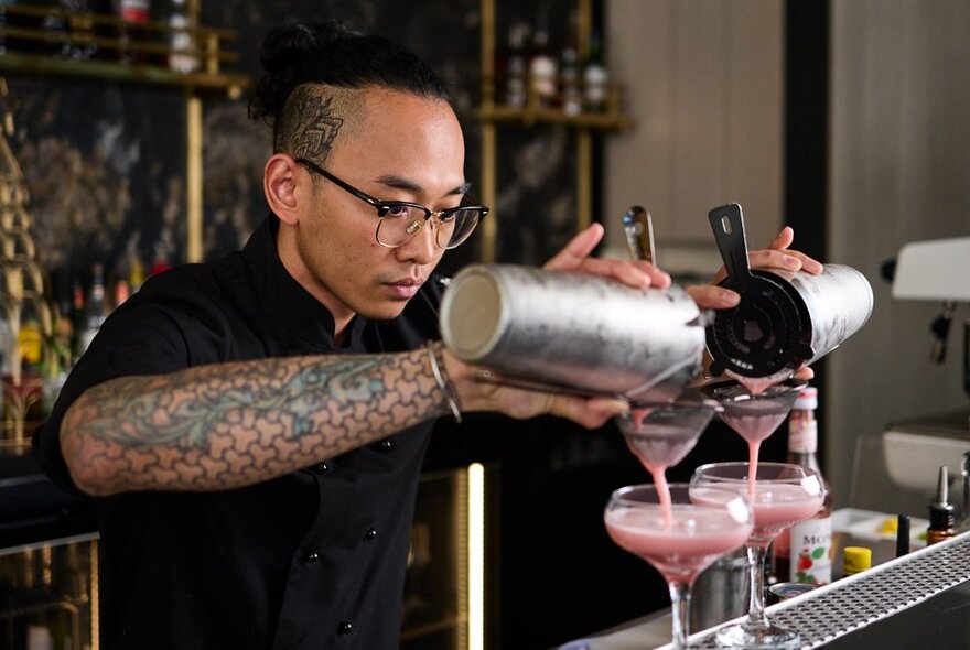 A bartender standing behind a bar and pouring two cocktails into martini glasses.