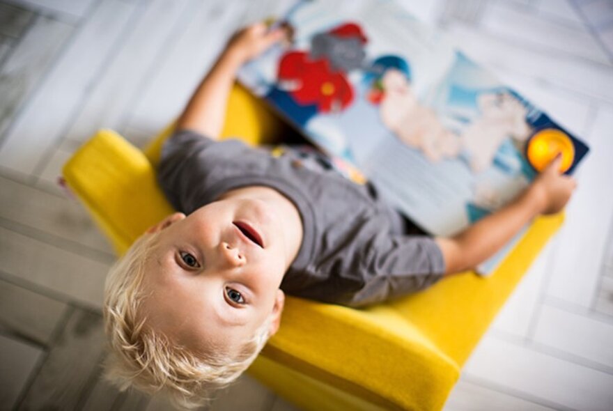 Looking directly down on a young child reading a picture book on a yellow chair, his head is back so he's looking directly at the camera.