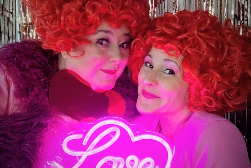 Two women wearing curly red wigs smiling beneath a pink neon sign with the word 'love' and silver glitter curtain.