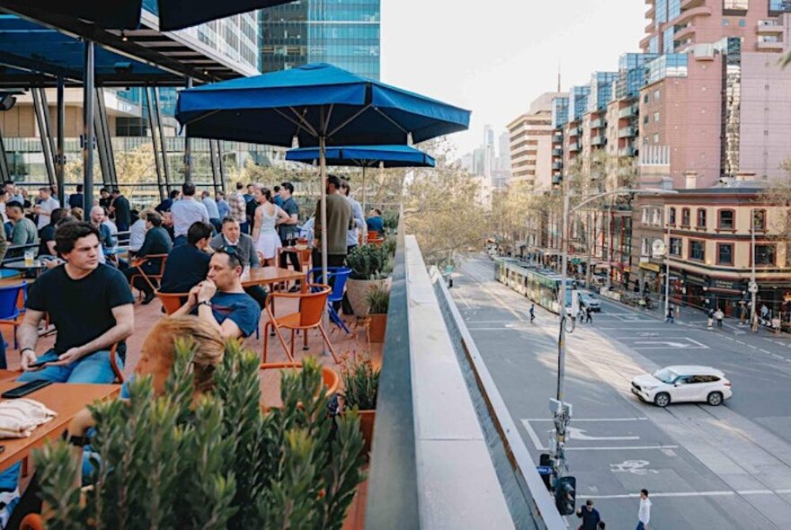 People drinking and relaxing at a crowded rooftop bar overlooking a long road in Melbourne. 