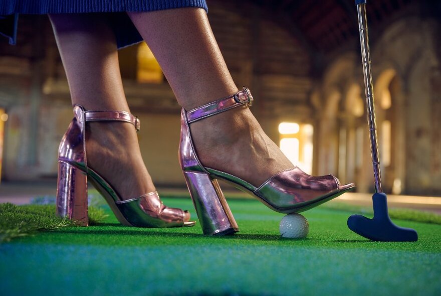 A pair of feet wearing pink metallic high heel shoes, on a green mini-golf putting runner of astroturf, with one foot gently pressing down on a golf ball, a golfing putter also in the frame. 