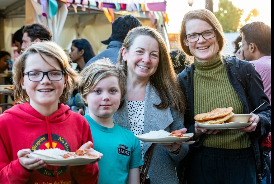 Two smiling adults and two children holding plates of Indian street food at the Festival of Diwali at Fed Square, other people in the background.