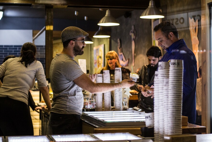 Customers being served gelati by a bearded man wearing a cap, standing at a counter with stacks of takeaway cups.