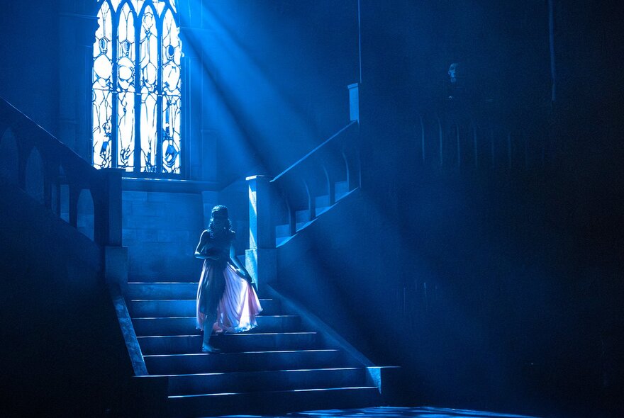 A figure on a dark staircase lit by tall Gothic stained-glass church windows.