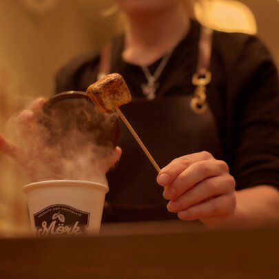 A person serving behind a counter, raising the lid on a takeaway cup of hot chocolate to add a marshmallow.