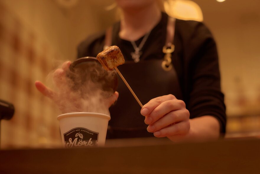 A person serving behind a counter, raising the lid on a takeaway cup of hot chocolate to add a marshmallow.